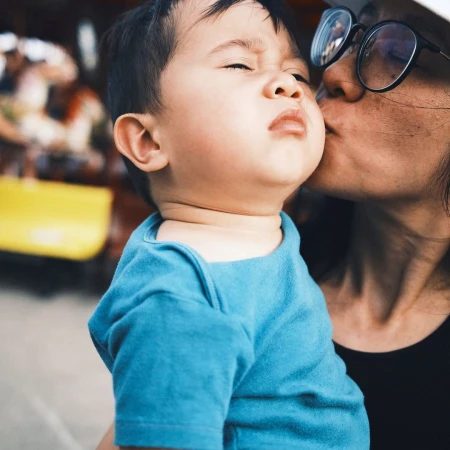 Close-up portrait of a woman wearing glasses kissing a toddler in a blue shirt on the cheek.