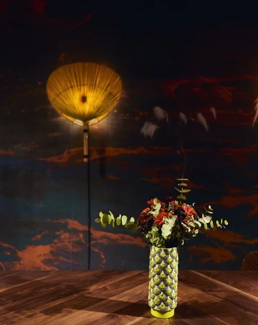 Floral arrangement in a patterned vase on a wood table, illuminated by a circular lamp against a cloud-patterned backdrop.  