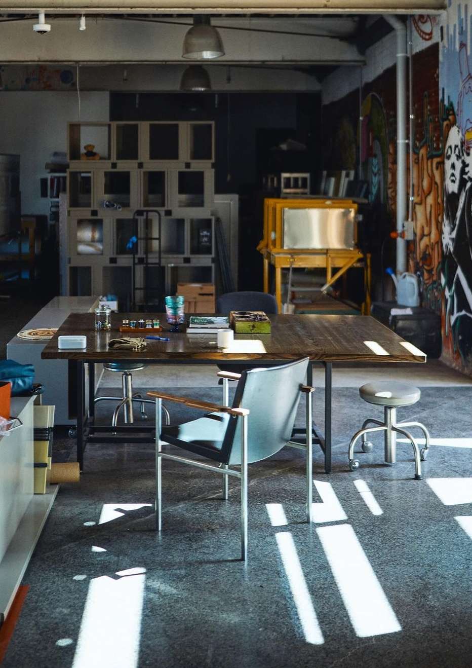 Photo studio interior with a dark wood table, metal chairs, and natural lighting casting shadows on the concrete floor.