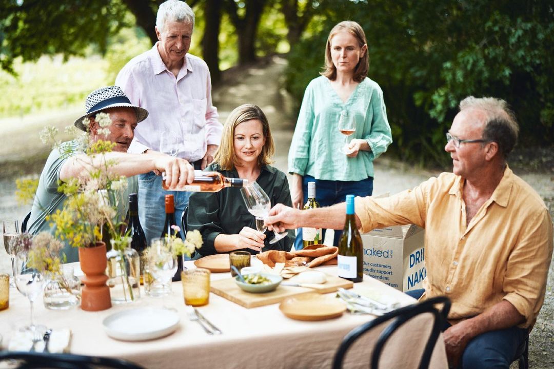 Outdoor wine tasting with five adults around a table with wine glasses, bottles, and food; greenery and trees in the background.