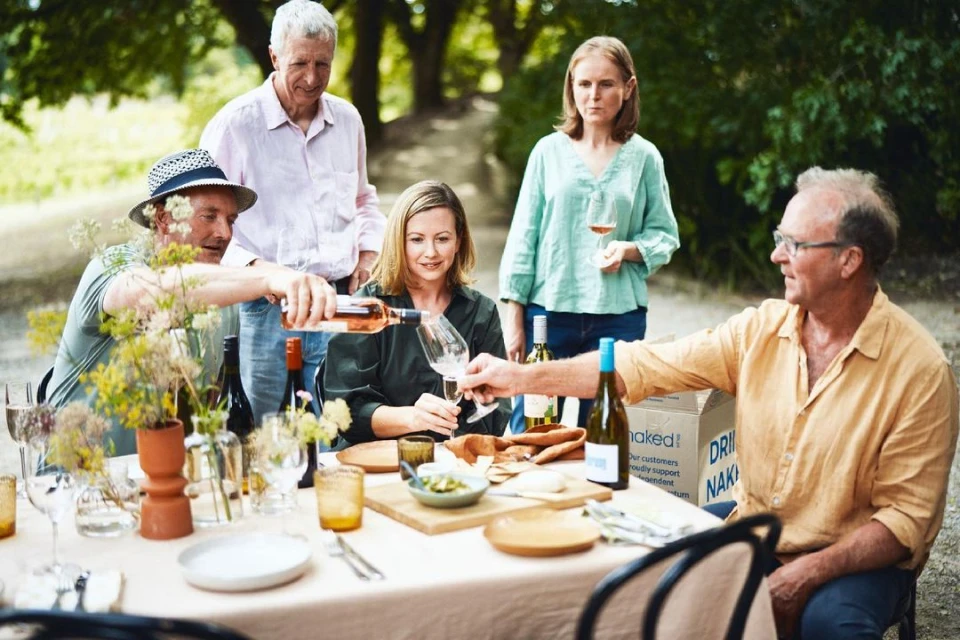 Outdoor wine tasting with five adults around a table with wine glasses, bottles, and food; greenery and trees in the background.