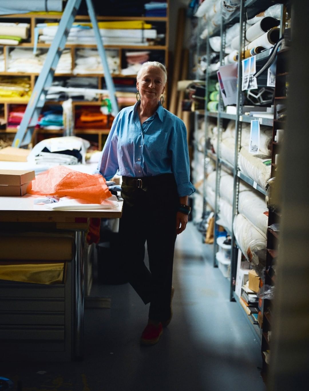 Eye-level medium shot of a smiling adult woman standing in a fabric storage room with shelves stocked with textiles and a table.