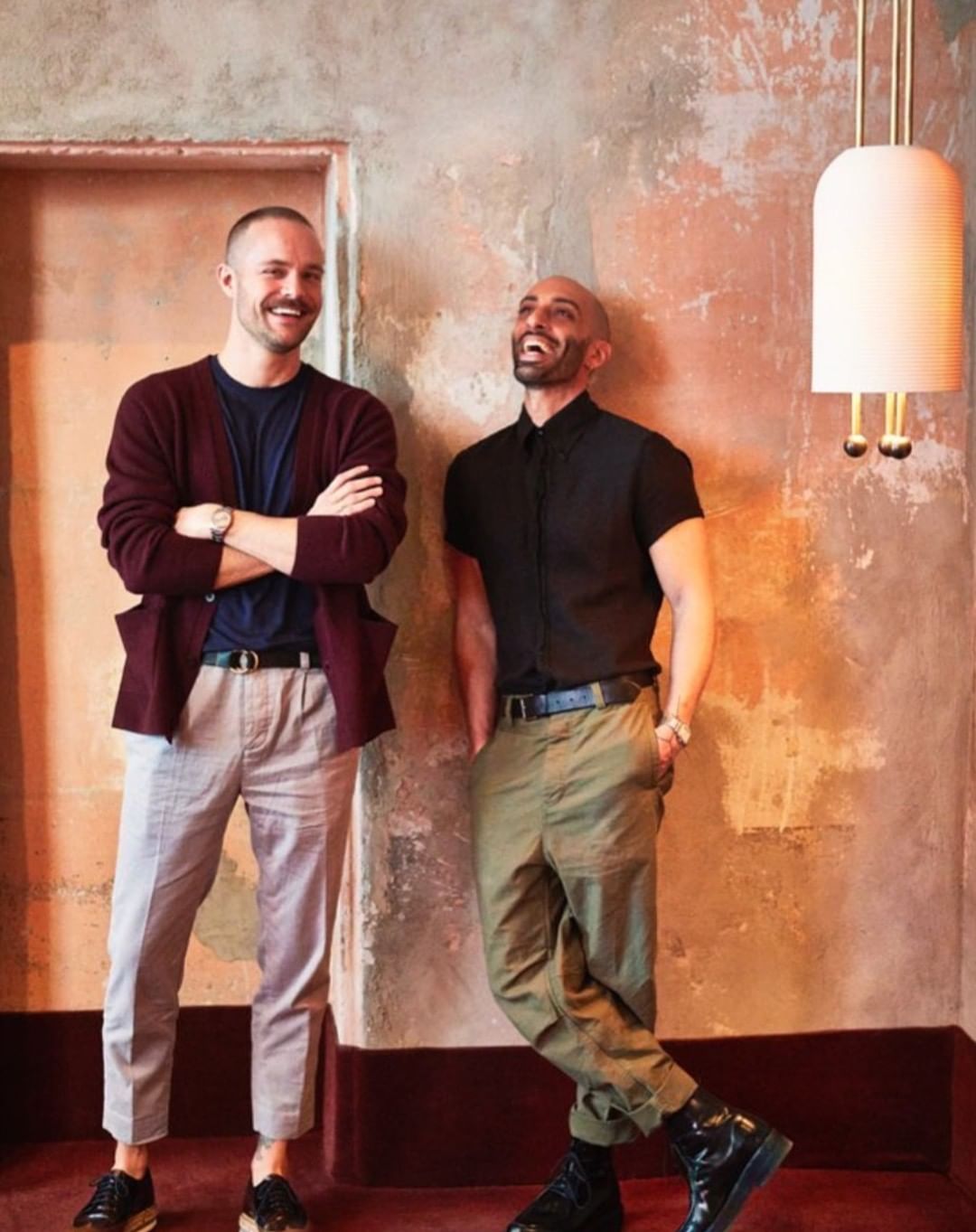 Two adult males pose indoors against a textured wall, one with arms crossed, the other laughing, in a lifestyle fashion shot.