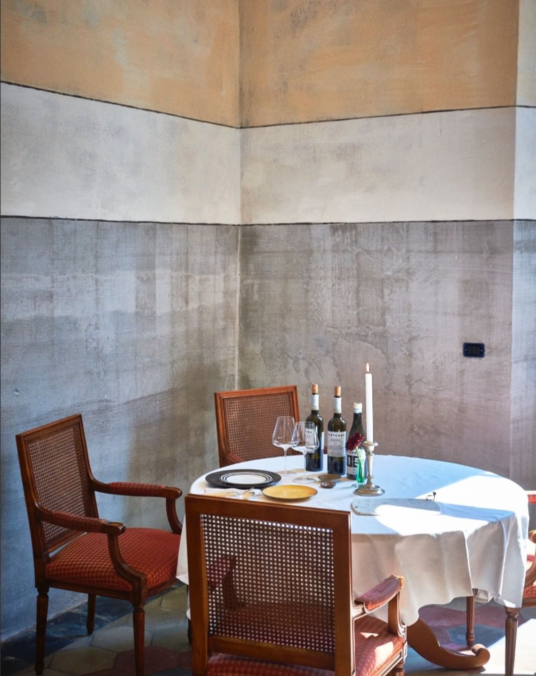 Dining room with four chairs, round table, tablecloth, wine bottles, wine glasses, plates, and candles indoors with natural light.