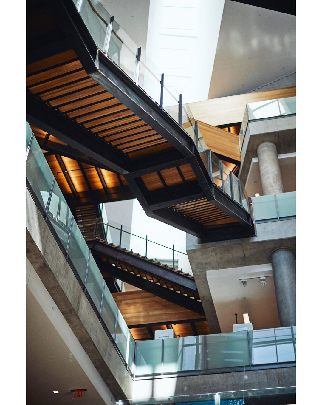 Architectural photo looking up at modern staircase and walkway system with glass railings, wooden treads, concrete supports, and natural lighting.