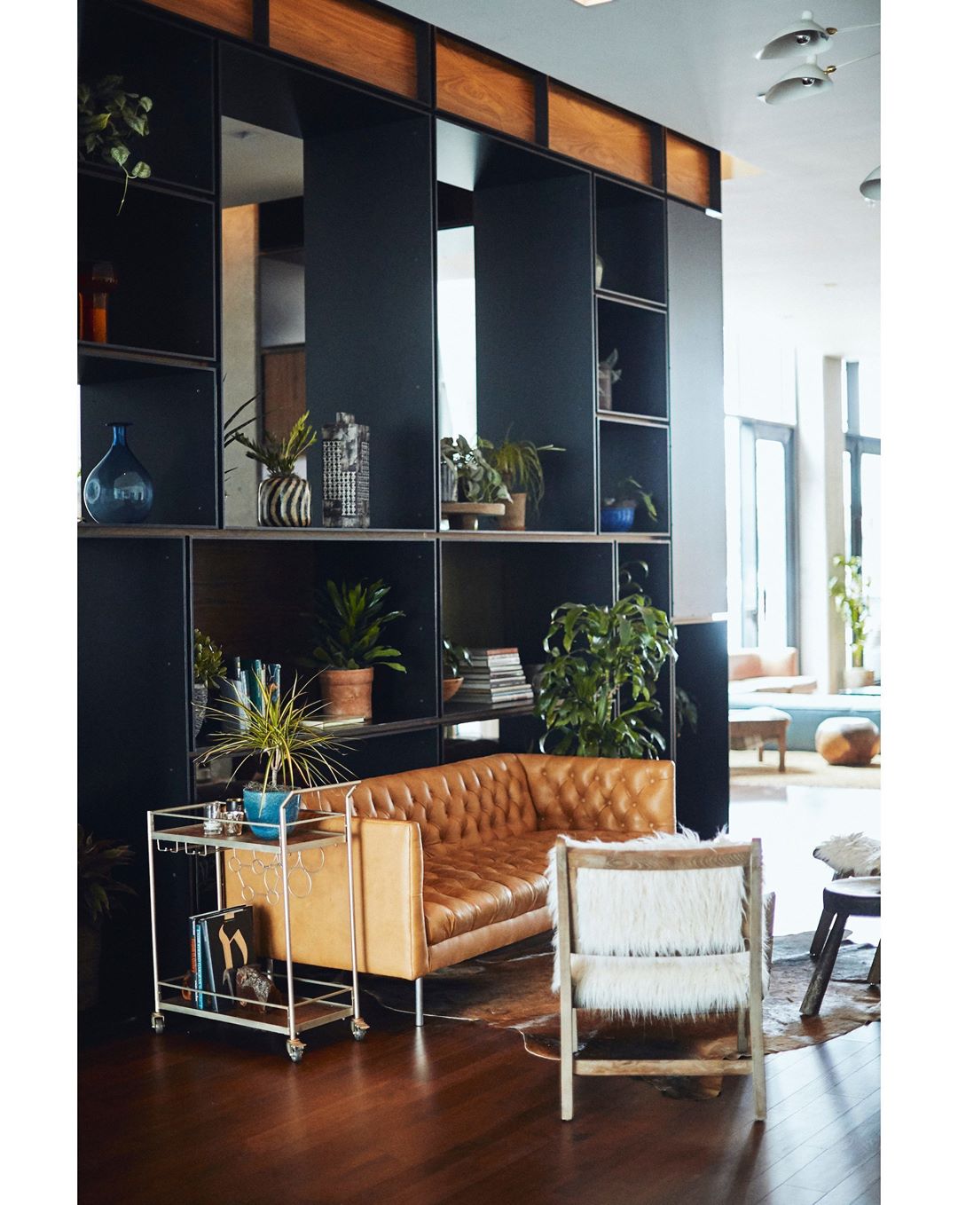 Living room interior showcasing a brown leather sofa, a white fur chair, a metal bar cart, and a black bookshelf.