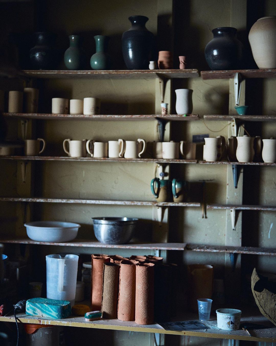 Various ceramic pottery and cups are arranged on shelves in a dimly lit room, creating a rustic and artistic studio scene.