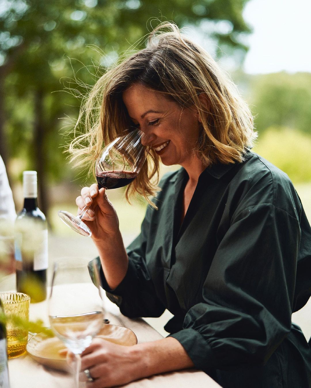 An adult woman smiling while holding a wine glass filled with red wine outdoors. Wine bottle and foliage are visible in the background.