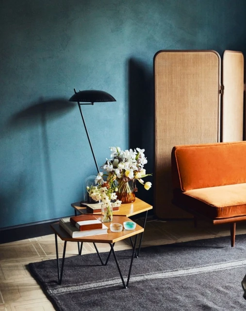 Living room interior featuring a modern orange couch, a pair of side tables with books and flowers, and a blue textured wall.