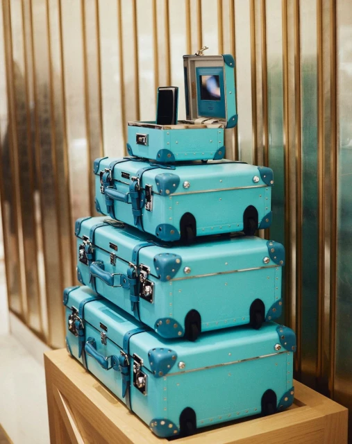 Stack of turquoise suitcases on a wooden base against a gold-toned metal bar background, conveying a sense of luxury travel.
