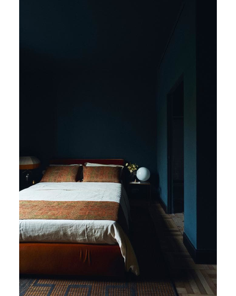 Bedroom interior with bed, nightstand, lamp. Features dark blue walls, patterned floor, and soft natural light in a residential setting.