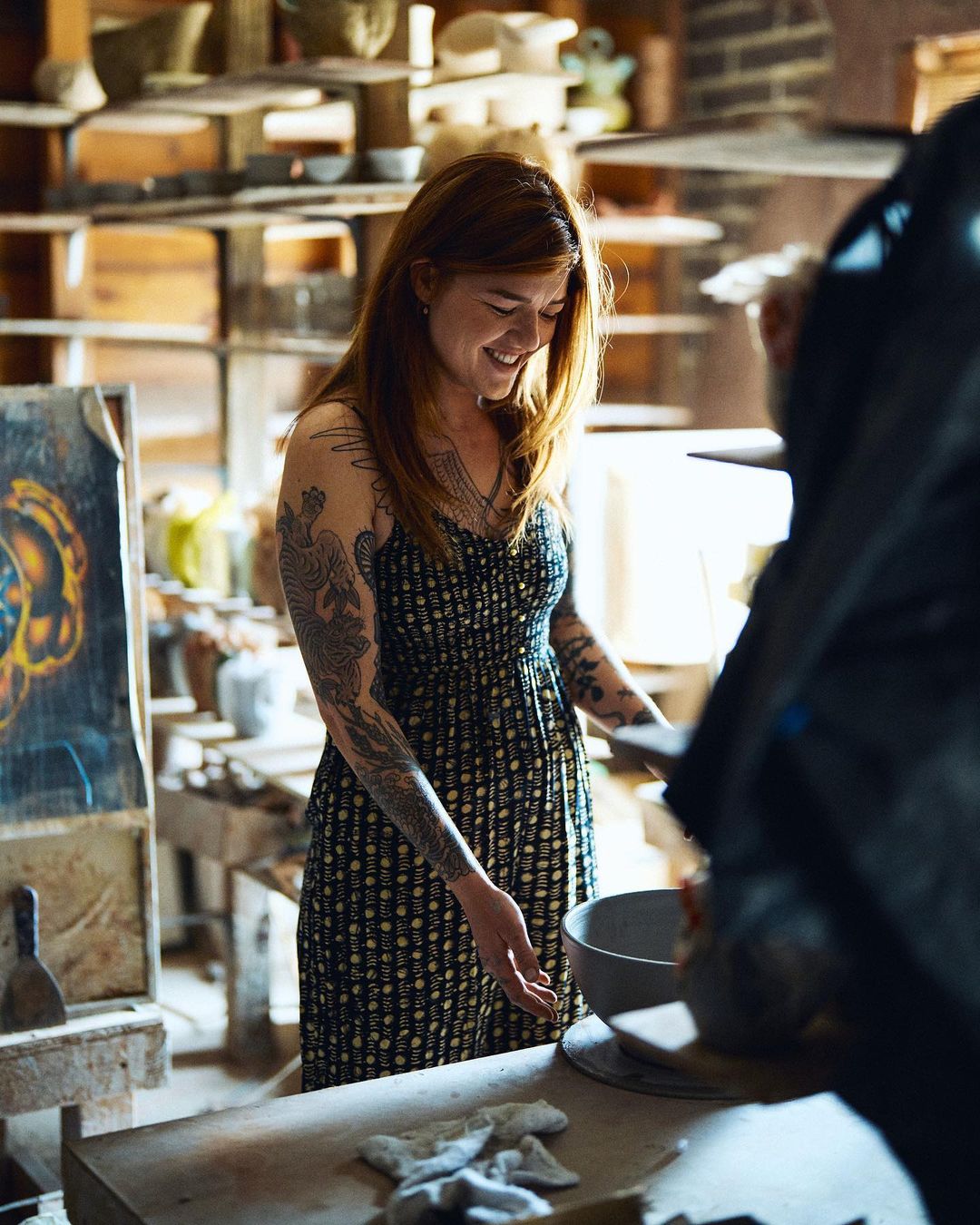 Smiling woman with tattoos in a dress stands inside a pottery studio with bowls, pottery tools, a table, and shelves in the background.