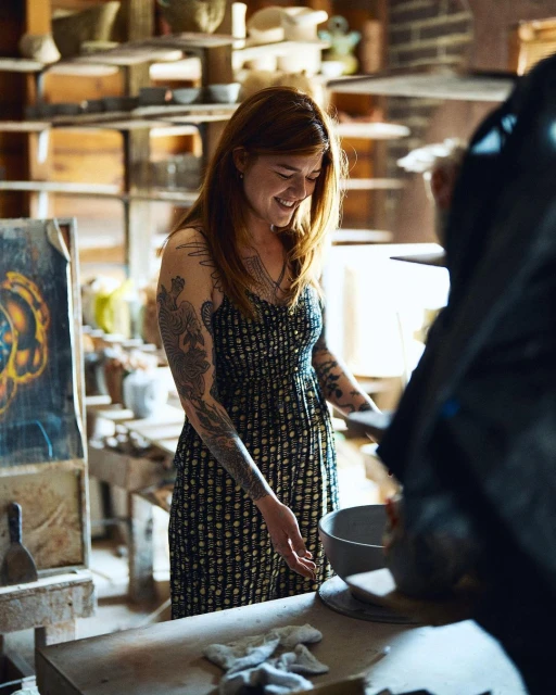 Smiling woman with tattoos in a dress stands inside a pottery studio with bowls, pottery tools, a table, and shelves in the background.