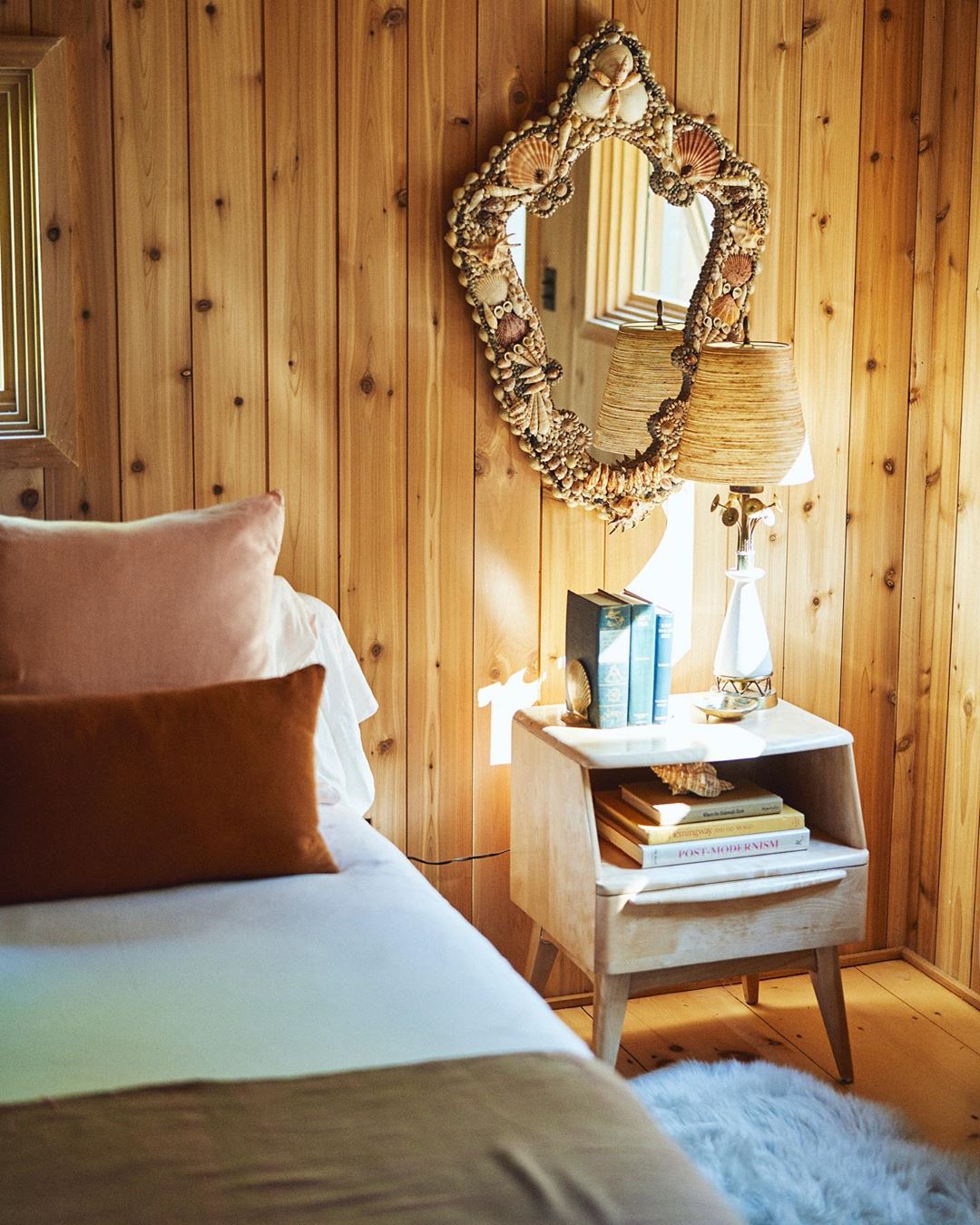 Interior view of a cozy bedroom with wood paneling, a seashell-framed mirror, a bed with neutral linens, and a vintage bedside table.