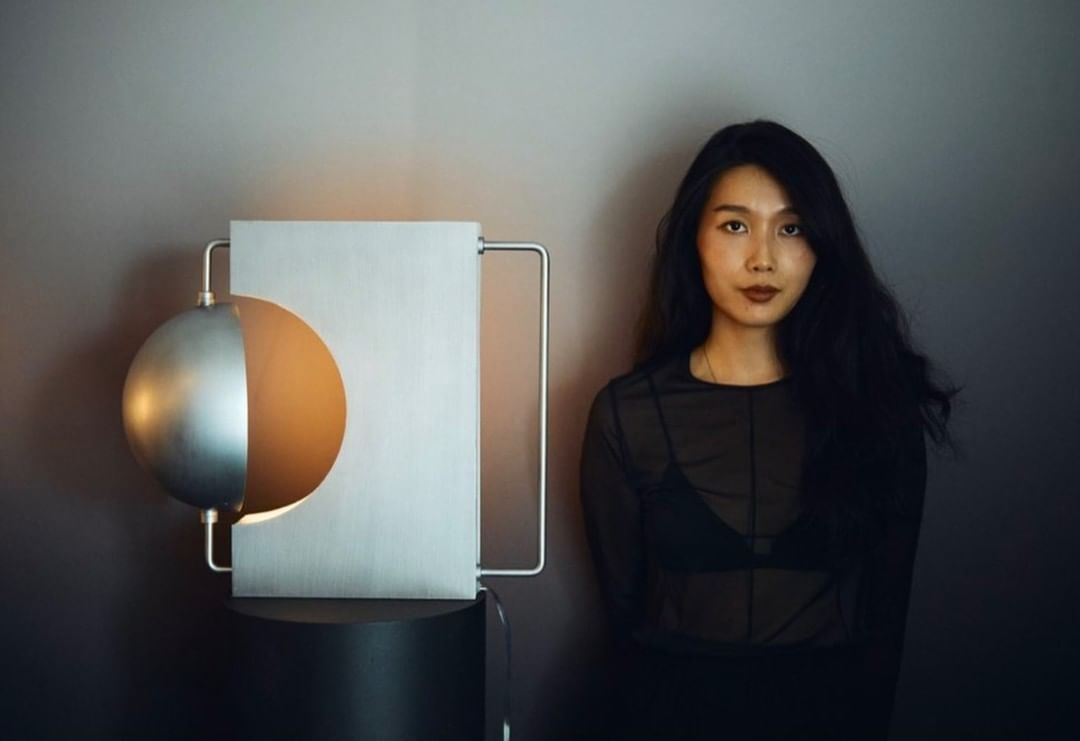 Portrait of an Asian woman with long dark hair and a sheer black top, posing next to a modern sculpture in a minimalist interior setting.