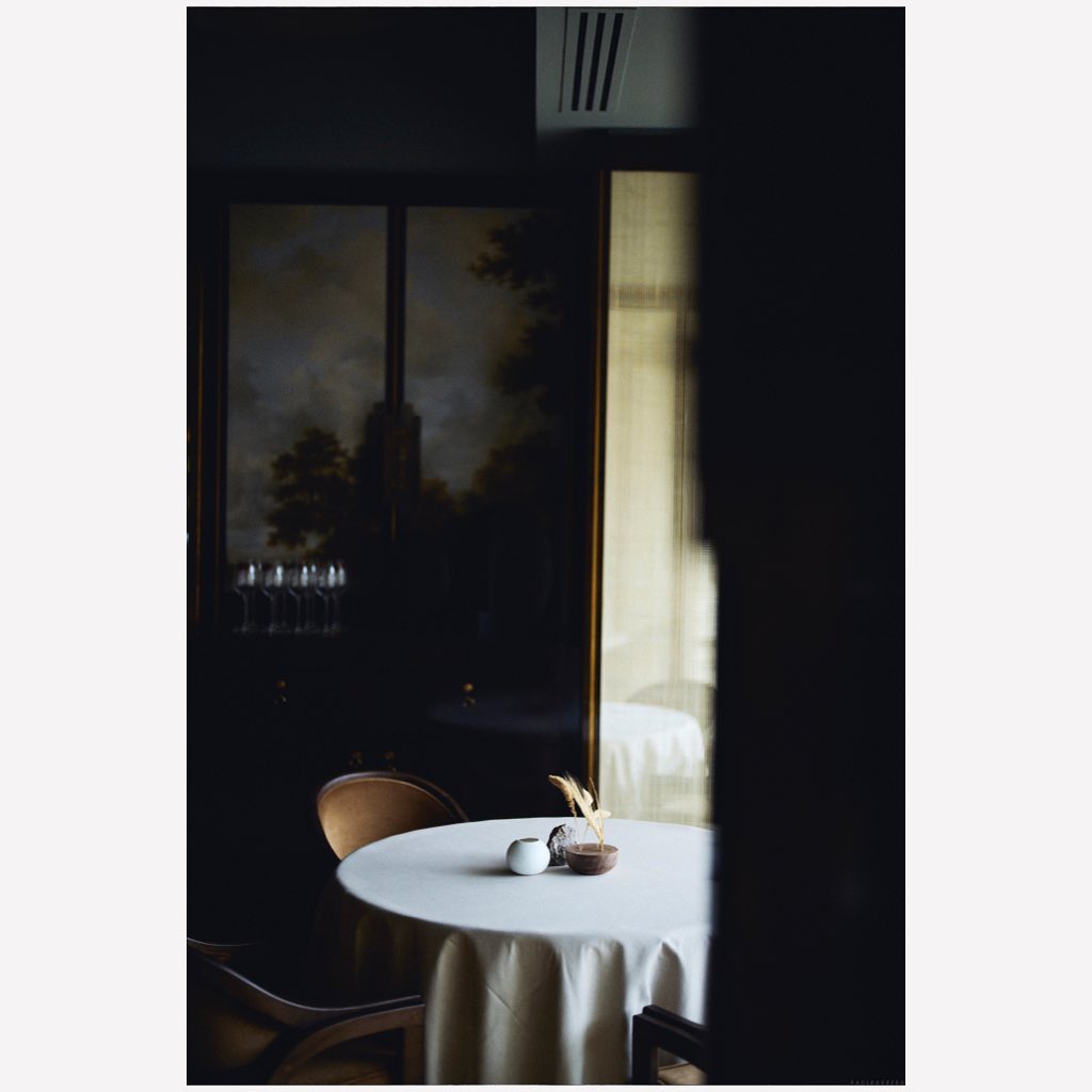 Elegant restaurant table setting with white tablecloth, glassware, and dark interior featuring a framed landscape painting.