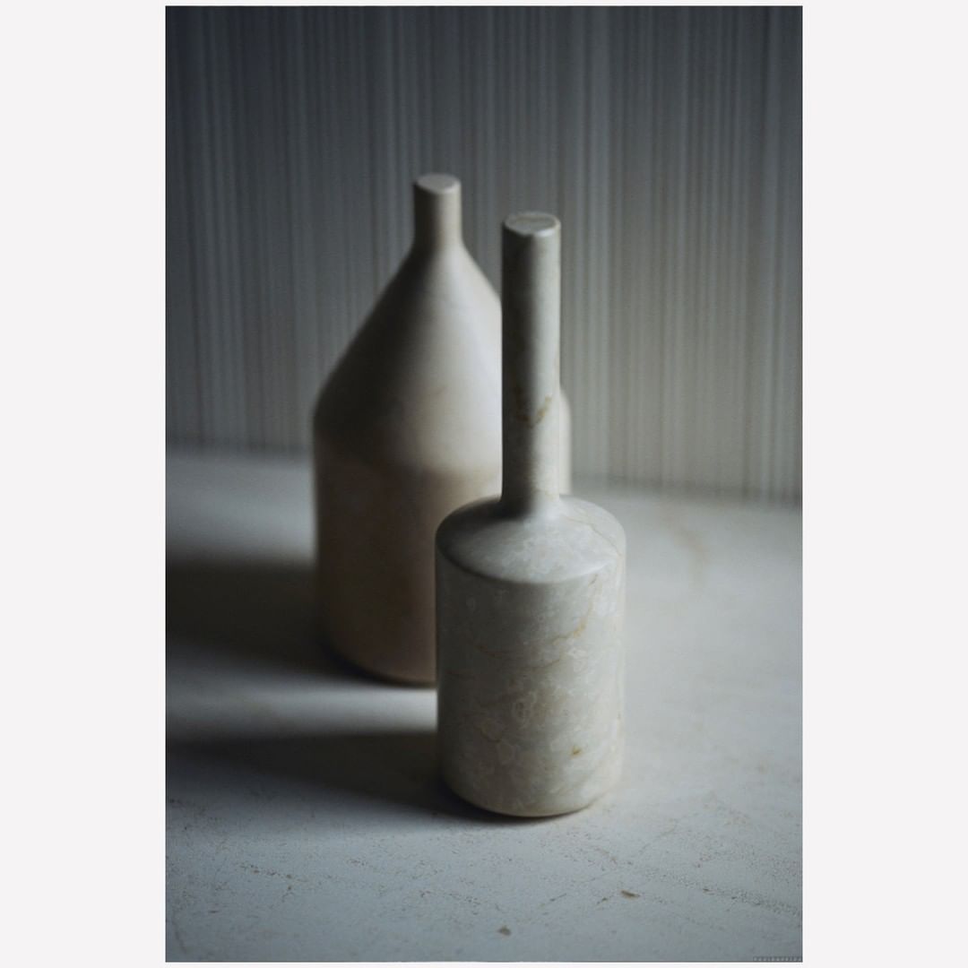 Still life of two minimalist beige vases on a white surface against a muted striped background with natural light and soft shadows.