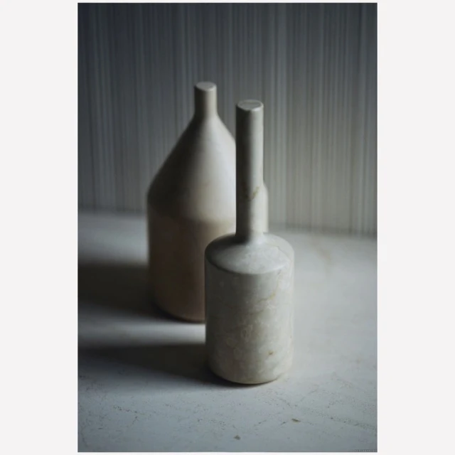 Still life of two minimalist beige vases on a white surface against a muted striped background with natural light and soft shadows.