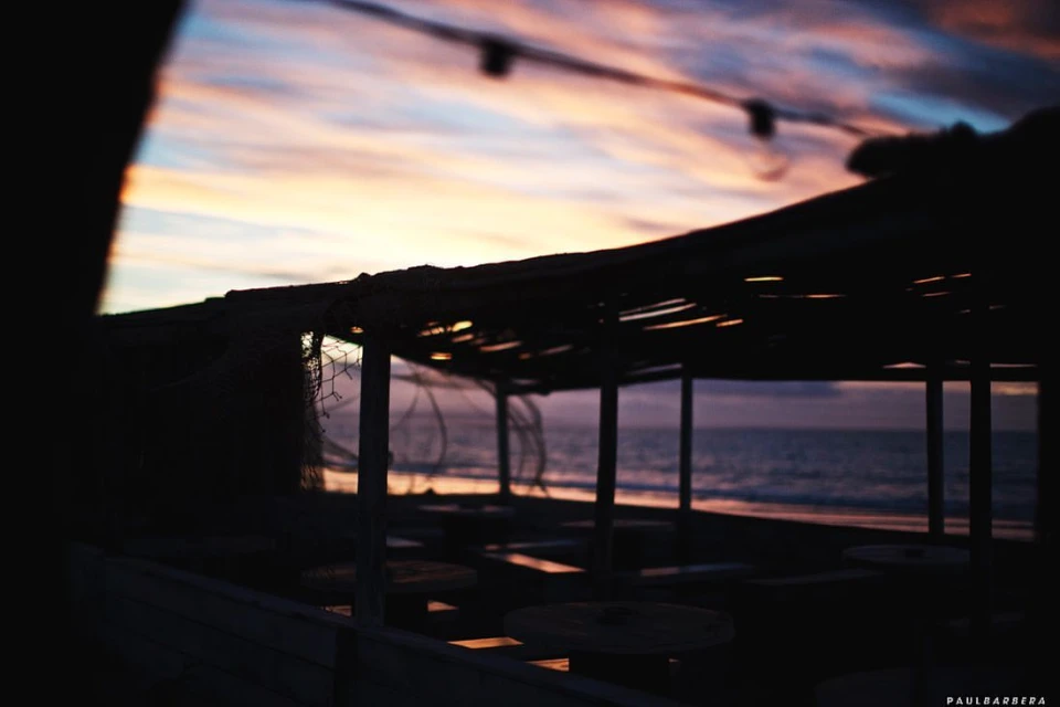 Coastal cafe at sunset features a thatched roof, columns, and tables silhouetted against a vibrant sky and calm ocean backdrop.