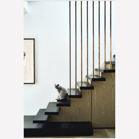 A grey cat sits on a black staircase, looking up with an alert expression, inside a modern interior with artwork on the wall.