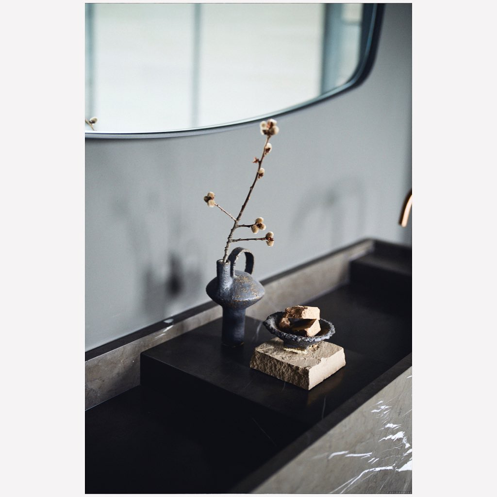 Modern bathroom interior with a black marble vanity, vase with floral branch, stone soap dish, and a neutral wall-mounted mirror.