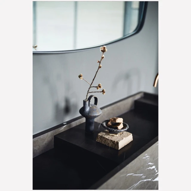 Modern bathroom interior with a black marble vanity, vase with floral branch, stone soap dish, and a neutral wall-mounted mirror.