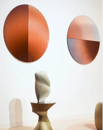 Modern art installation featuring textured circular wall hangings and a sculptural vase displayed on a metallic pedestal in a gallery setting.