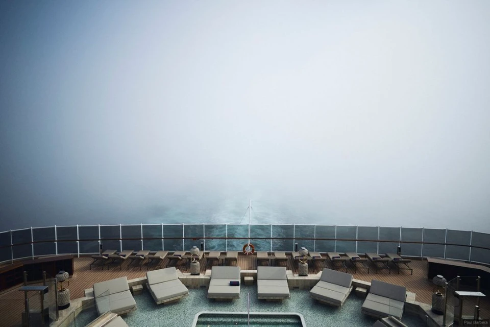 Aerial view of a cruise ship deck featuring lounge chairs and a pool, overlooking a foggy ocean with visible ship wake.