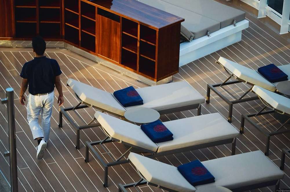 Man walks across deck with lounge chairs and wood flooring. A cabinet with shelving is in the background. Cruise vacation scene.