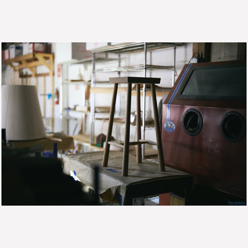 Wooden stool resting on a covered workbench inside a workshop with metal shelving and a red sandblasting machine.