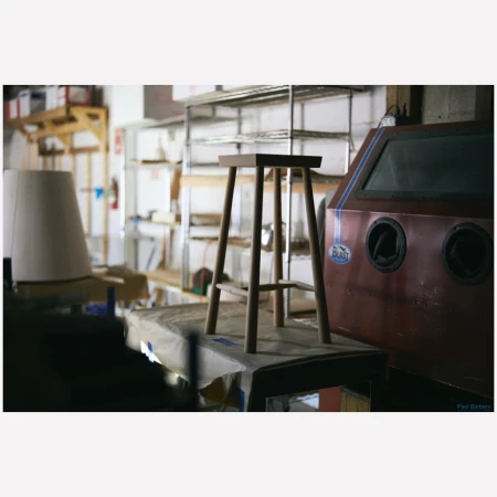 Wooden stool resting on a covered workbench inside a workshop with metal shelving and a red sandblasting machine.