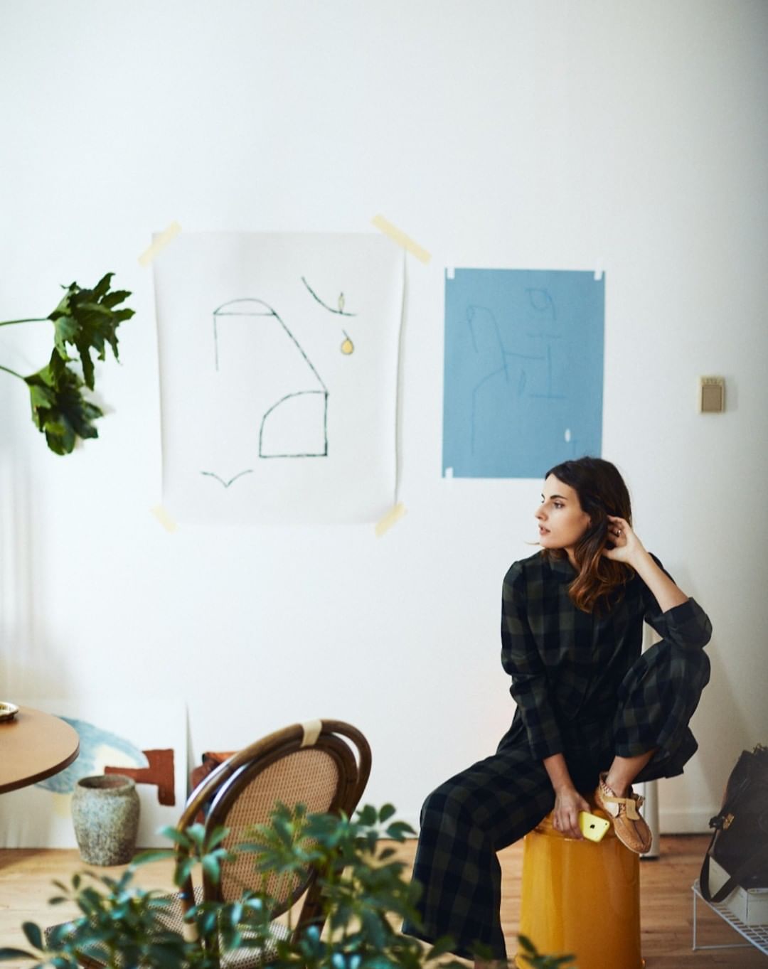 Adult woman wearing a plaid outfit seated on a yellow stool in a living room with modern art and plants; natural daylight.