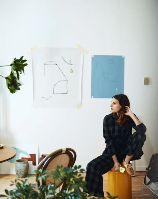 Adult woman wearing a plaid outfit seated on a yellow stool in a living room with modern art and plants; natural daylight.