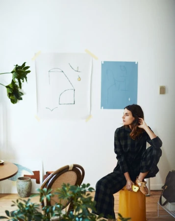 Adult woman wearing a plaid outfit seated on a yellow stool in a living room with modern art and plants; natural daylight.