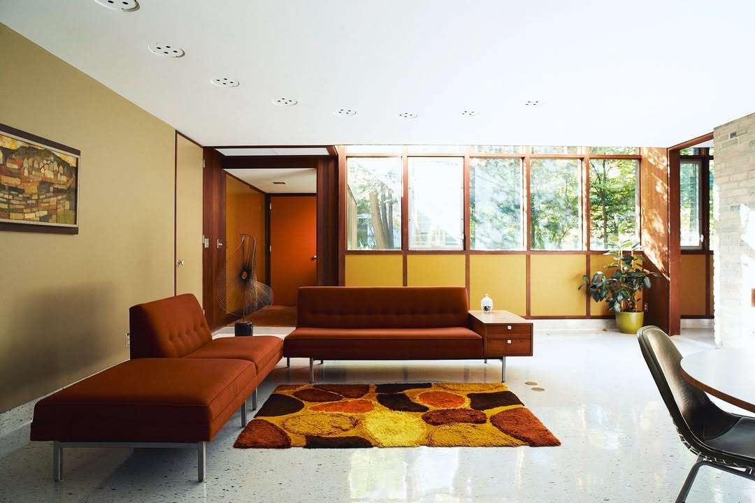 Living room interior with brown sectional sofa, rug with brown and orange geometric patterns, and large windows with a view of trees.