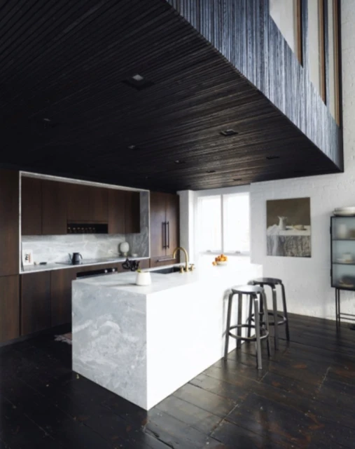 Modern kitchen interior featuring a marble island with stools, dark wood cabinets, and a high ceiling with unique architectural details.