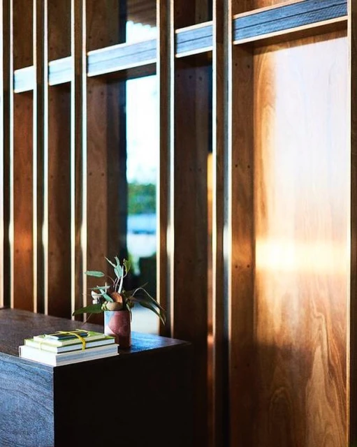 Interior shot of a dark wood table with stacked books and a potted plant in front of a wood paneled wall and window.