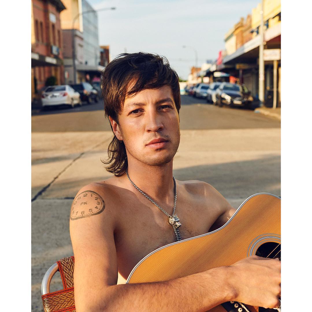 Adult male musician poses outdoors with guitar on an urban street with cars and buildings visible in the background on a sunny day.