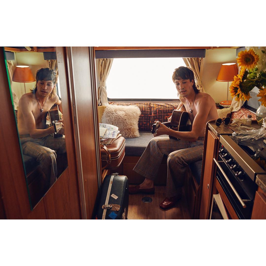 Man plays guitar inside camper van next to mirror and sunflowers, sitting by a window with luggage and a cooking range visible.