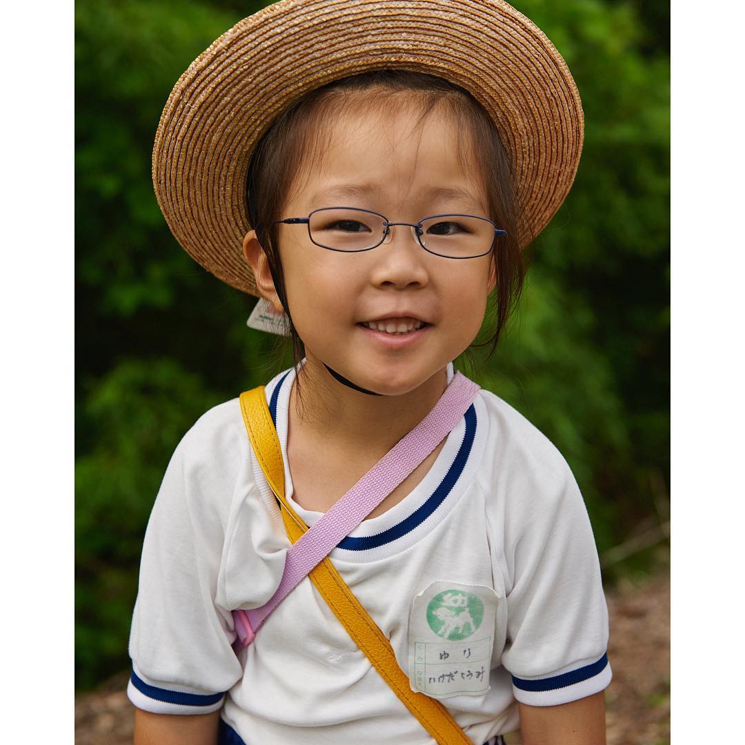 Young child wearing a straw hat, glasses, and a white t-shirt with a colored strap across the chest outdoors.