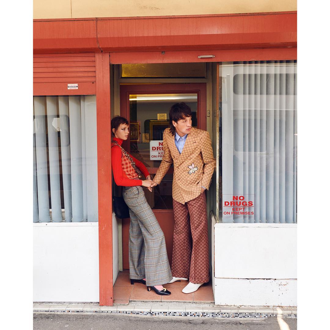 Stylish couple poses in doorway wearing patterned retro fashion. A woman in red turtleneck, and a man in patterned jacket hold hands.