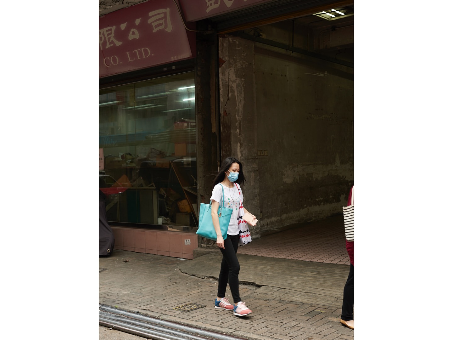 Woman in face mask walks on sidewalk near building with Chinese signage, carrying blue tote bag in an urban setting during the daytime.