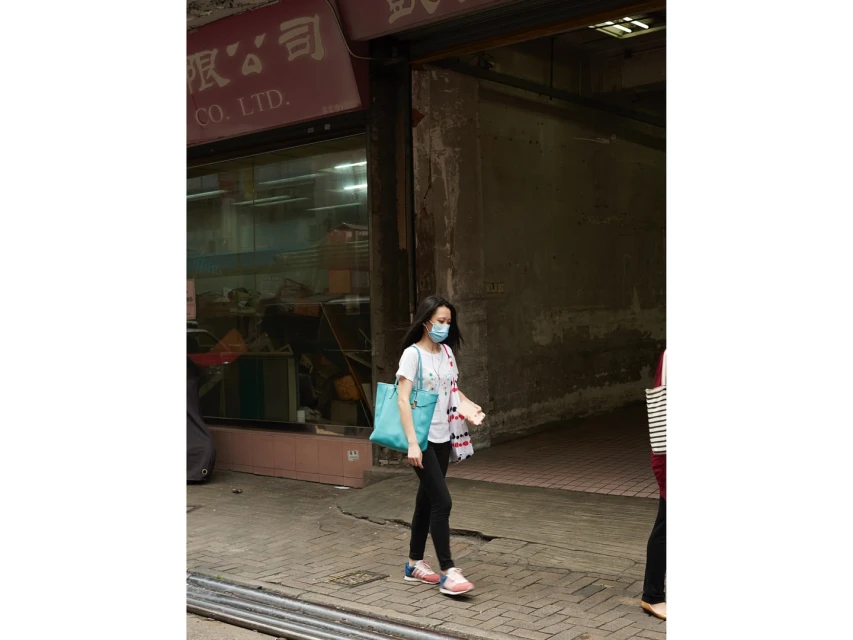 Woman in face mask walks on sidewalk near building with Chinese signage, carrying blue tote bag in an urban setting during the daytime.
