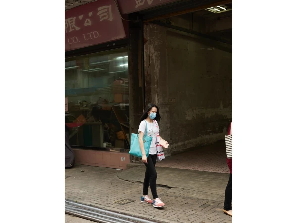 Woman in face mask walks on sidewalk near building with Chinese signage, carrying blue tote bag in an urban setting during the daytime.