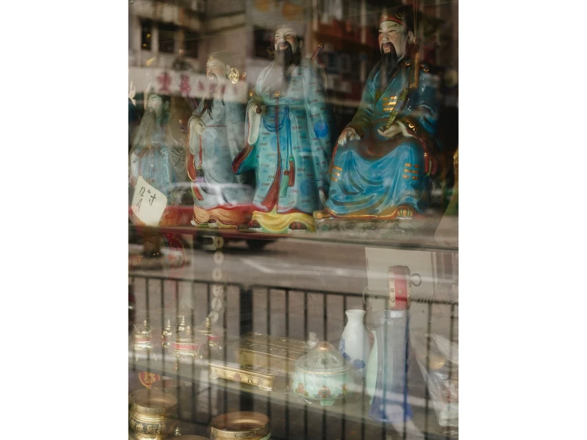 Ornate Asian figurines displayed in a shop window, reflecting street view. Gold and blue colors, various ritual vessels also visible.