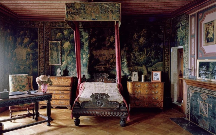 Ornate four-poster bed in historic interior with tapestry-covered walls, wooden desk and furniture, and parquet wood flooring.