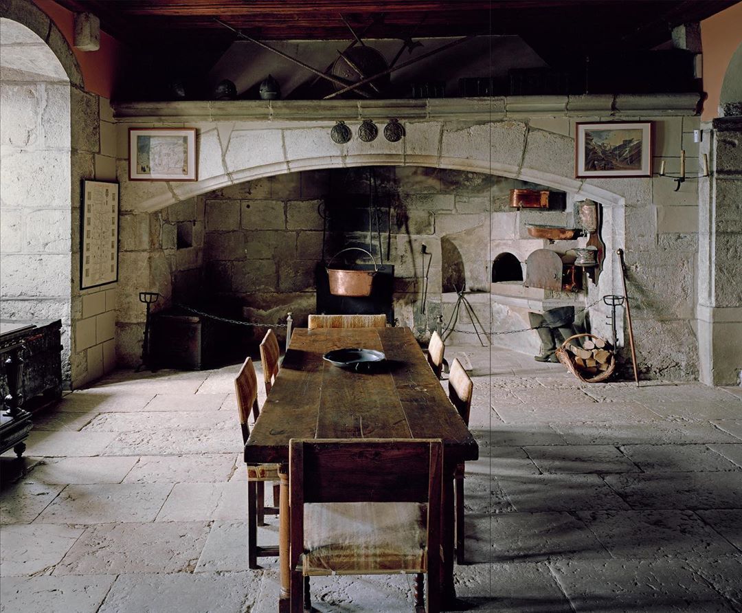 Rustic kitchen interior featuring a long wooden table, stone fireplace, and vintage decor, evoking a historic or medieval ambiance.