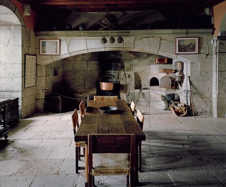 Rustic kitchen interior featuring a long wooden table, stone fireplace, and vintage decor, evoking a historic or medieval ambiance.