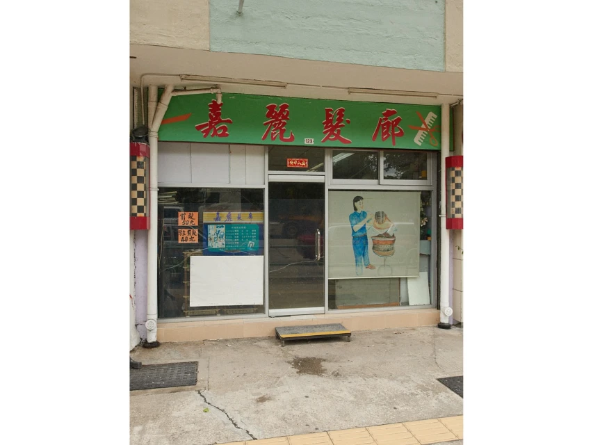 Exterior of a small, green-painted building in daylight with a large sign and window displaying a woman washing hair.