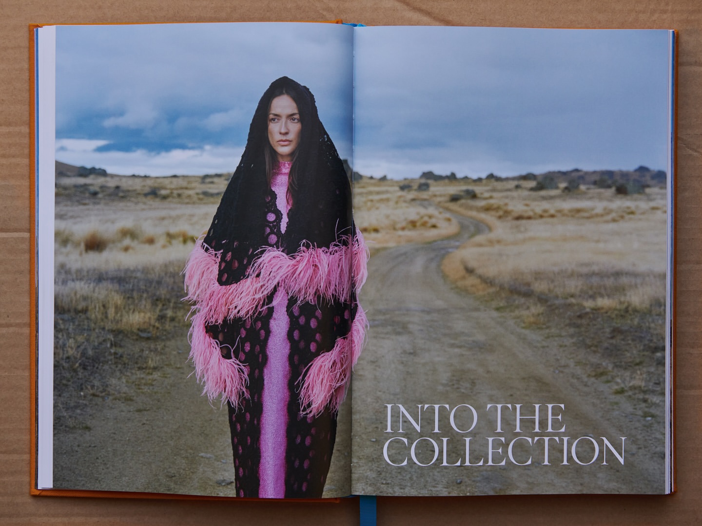 Fashion editorial shot on an open book page with a model wearing a fringed shawl in an arid landscape under a cloudy sky.